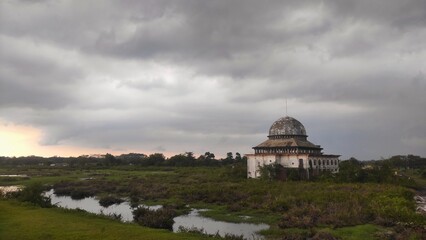 Fototapeta premium An abandoned mosque stands silently under a cloudy sky, surrounded by overgrown vegetation and small water pools, creating a moody and atmospheric scene.