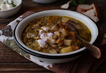 Vegetarian potato and bean soup served in rustic enameled bowl on wooden table. Sliced bread on the side