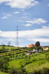 Un paisaje rural con colinas verdes, casas dispersas y una gran torre de alta tensi&oacute;n bajo un cielo azul con nubes.

