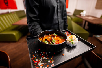 A waiter serves a bowl of flavorful Asian cuisine, featuring vibrant vegetables and herbs, accompanied by a side of sushi on a black tray in a modern restaurant setting.