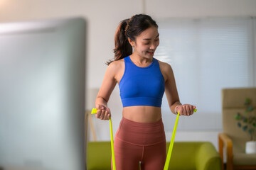 Young asian woman doing fitness exercises at home using a resistance band and watching online...