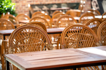 Empty wooden tables and wicker chairs on an outdoor patio. Sunlight illuminates the rustic setting, perfect for cafe, restaurant, or summer themes
