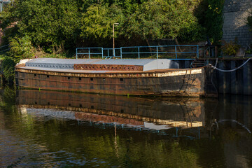 A weathered barge sits still on a canal waterway, its reflection mirrored in the water. Surrounding...