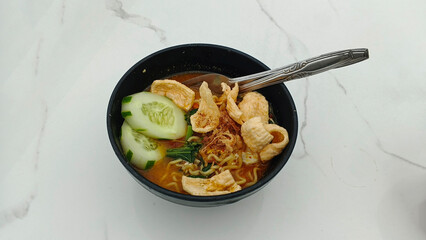 The bowl contains cooked noodles in a broth, garnished with slices of cucumber, fried shallots, and crispy crackers.