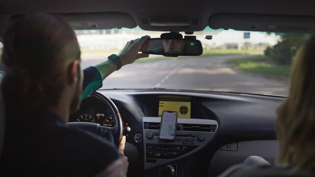 Caucasian male driver adjusting rearview mirror while steering modern car with female passenger sitting beside him. Vehicle moving through suburban roads on sunny day. Driver ensuring safe journey.