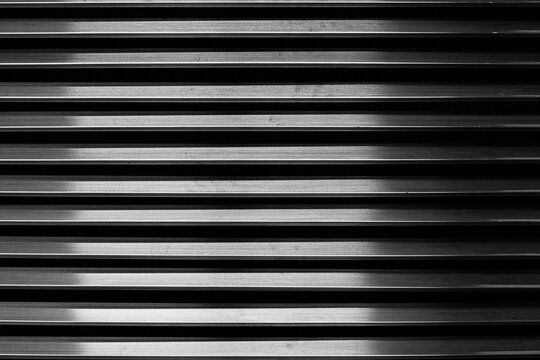 The parallel metal bars of the ventilation grille create a repeating pattern, enhanced by the play of light and shadow, adding depth and texture to the metal surface. Metal grille background.