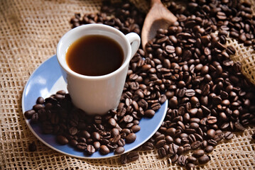 Freshly brewed coffee inside a coffee cup and roasted coffee beans on a rustic surface with a wooden spoon. Top view.