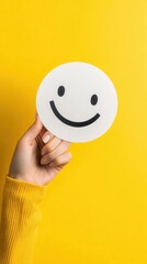 A hand holds a paper cut-out with a smiley face against a bright yellow background, symbolizing positivity and happiness.