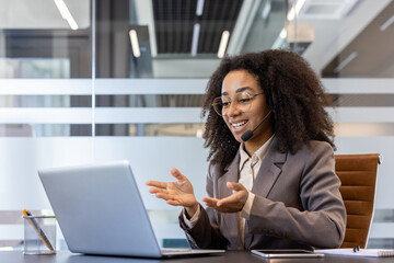 Smiling young African American woman in suit and headset sitting at desk in office and gesturing with hands talking on video call on laptop