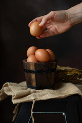 Hand holding brown egg over the round wooden bowl full of brown eggs, perfect for Easter on dark background 
