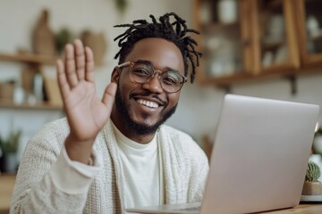 Obraz premium A smiling man with dreadlocks and glasses waves at his laptop screen while sitting in a warmly lit room. The image represents virtual communication, remote work, and online meetings.