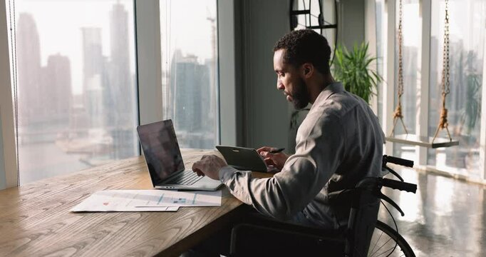 Serious young African American businessman with disability working on project in office, using laptop, sitting in wheelchair at large window, typing, writing notes, analyzing electronic information