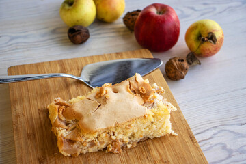 Piece of traditional thanksgiving apple cinnamon pie on the wooden cutting board on the white table, surrounded by fresh apples and walnuts. Autumn dessert. Closeup. Copy space.