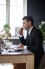 A young man in a black suit enjoys a cup of coffee while sitting at a desk with a laptop. The bright office features plants, a calendar, and a relaxed, professional atmosphere.