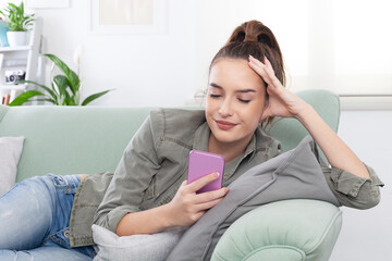 Young relaxed smiling woman lying on a cozy couch in a casual outfit, holding a smartphone and browsing messages, social media, and digital content