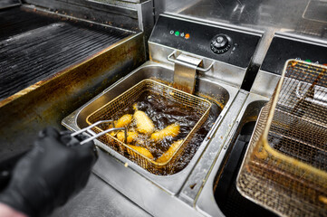 A chef's hand using tongs to remove golden-brown fried food from a deep fryer. The kitchen equipment is stainless steel, with bubbling oil visible in the fryer basket.