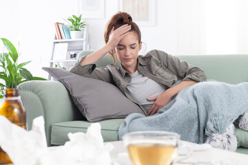 Young woman lies on the couch, holding her head and stomach in pain, suffering from a headache and abdominal discomfort, due to period cramps or flu, trying to relieve with tea and medication pills