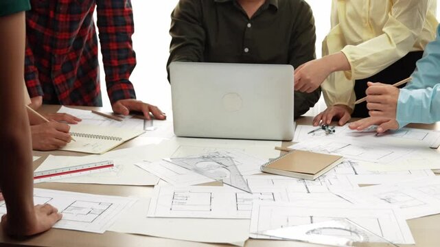 Architect and safety engineer discussion about building blueprint displaying on laptop on table with architectural document and wooden block scatter around at modern office. Closeup. Delineation.