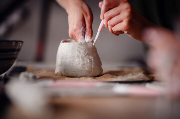 Close-up of hands meticulously detailing a clay pottery piece using a sculpting tool. The image...