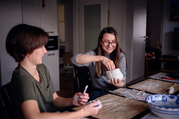 Two women engaged in a clay sculpting project, conversing and sharing a creative moment. The image captures collaboration and artistic expression in a relaxed setting.