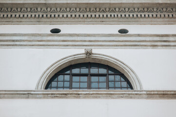 An old semicircular window with black iron bars in a frame on a light gray wall. Attic. Jesuit Church in Lviv.