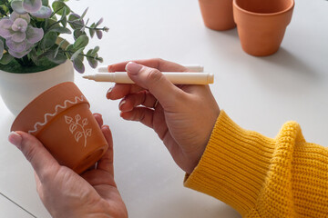 Caucasian woman decorating a terracotta pot with a white marker in a bright, cozy setting