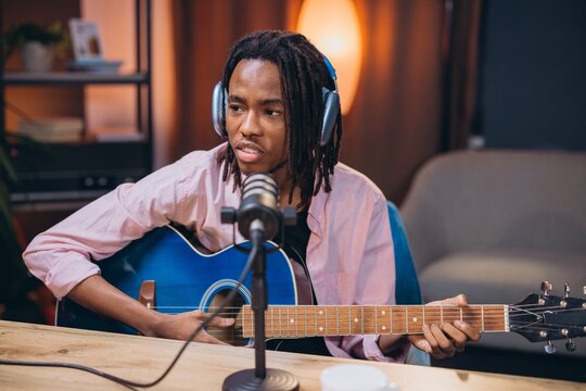 Young African American man with dreadlocks wearing headphones playing an acoustic guitar and recording a podcast in a home studio
