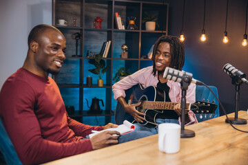 Two African American men recording a podcast in a home studio, one playing guitar and the other...