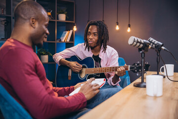 African American musician performing acoustic guitar during podcast recording, sound engineer noting details in home studio setting