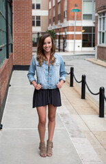 Beautiful young woman poses wearing short black dress and denim jacket standing in front of brick building in city in middle of sidewalk