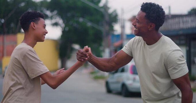 Father of African descent bonding with teenage son through a heartfelt embrace and a joyful handshake in a relaxed urban street, celebrating trust, connection, and family unity