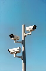 Three white security cameras mounted on gray pole against clear blue sky. Urban tech equipment for safety and security. Modern surveillance system. City tech infrastructure. Public safety in action.