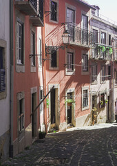 Naklejka premium A charming cobbled street lined with pastel-colored buildings, wrought-iron balconies, and potted plants. Sunlight casts soft shadows, enhancing the quaint European atmosphere.