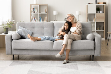 Affectionate aged grandmother hugging caressing dear little granddaughter on large comfy sofa....