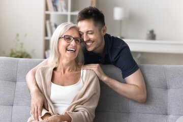 Fototapeta premium Visiting mom. Affectionate young adult son hug smiling elder aged mother sitting on couch with warmth care lean head to her with closed eyes sincere smile feel gratitude to single parent grown him up