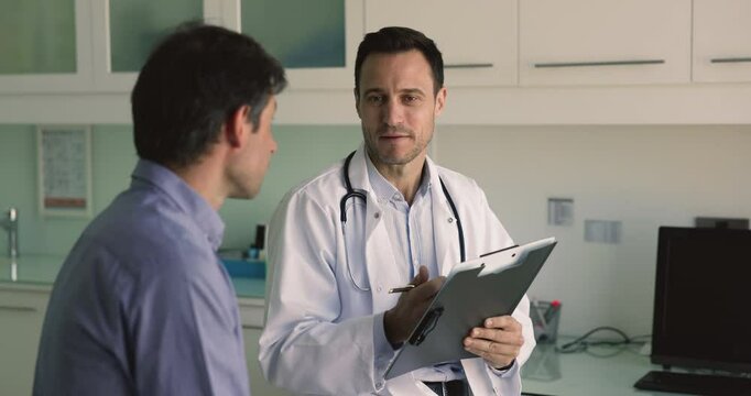 Positive male Latin doctor talking to patient on appointment in clinic, filling up survey, writing notes, keeping records, asking about healthcare complaints, sickness symptoms, smiling, speaking