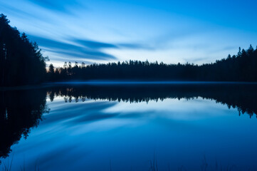 A scenic and calm lake view at night with mist and reflected forest on the surface in Finland
