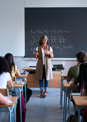 Vertical. A teacher stands at the front of a classroom, giving a lesson to her students. The scene...