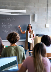 Vertical. View from behind a group of multiracial students taking notes in a classroom. The teacher...