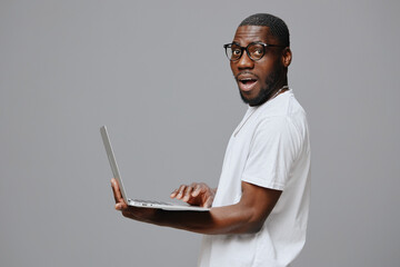 Excited young man using laptop in casual white t shirt, showcasing enthusiasm while working Grey background enhances the focus on the model s expression and outfit