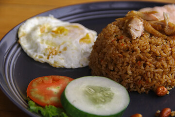 Fried rice with fried egg, peanuts, anchovies, cucumber, tomatoes and crackers on a plate on a wooden background. Indonesian food