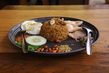 Fried rice with fried egg, peanuts, anchovies, cucumber, tomatoes and crackers on a plate on a wooden background. Indonesian food