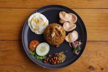Fried rice with fried egg, peanuts, anchovies, cucumber, tomatoes and crackers on a plate on a wooden background. Indonesian food