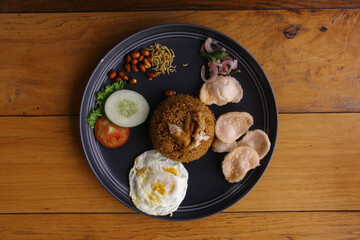 Fried rice with fried egg, peanuts, anchovies, cucumber, tomatoes and crackers on a plate on a wooden background. Indonesian food