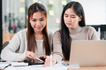 Two young women working together on a project using a tablet and laptop in a modern office setting.
