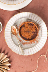Cocoa dessert on a white plate, dusted with cocoa powder, a golden spoon beside it, set on a brown surface with additional cocoa scattered around. Top view