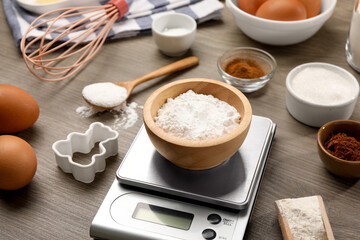 Kitchen scale with bowl of baking powder, eggs, sugar and cocoa on wooden table, closeup