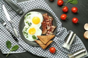 Tasty fried eggs, bacon and pieces of toast served on black table, flat lay