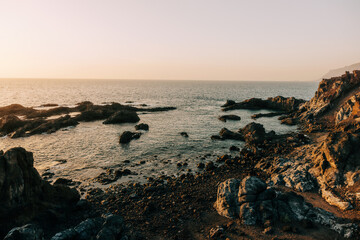 Rocky coastal landscape with calm sea and soft golden light during sunset.