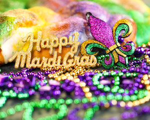 Happy Mardi Gras text in gold glitter and a king cake with yellow, green, and purple sprinkles surrounded by Mardi Gras beads and a glittering fleur de lis. © Leigh Prather